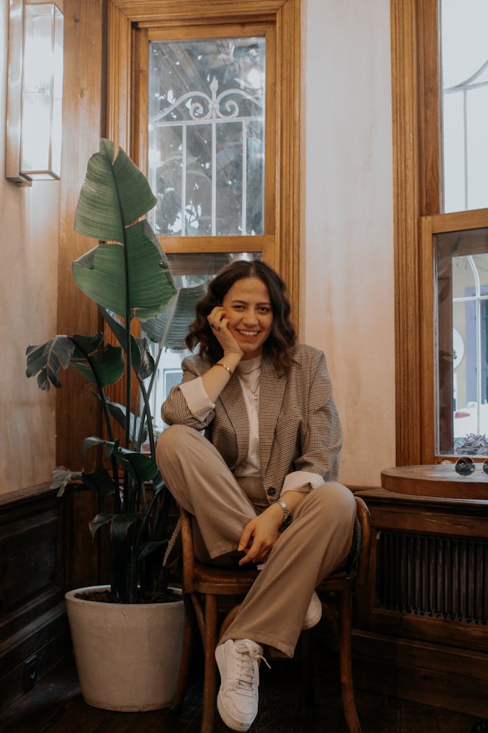 Elegant woman in a blazer sits on a chair with potted plant in cozy indoor setting.